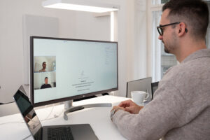 A man sits at a desk participating in a video call on a large computer monitor. The screen shows two people in a video meeting and a digital document with a signature section. A laptop, coffee cup, and notepad are on the desk in a bright, modern office setting.